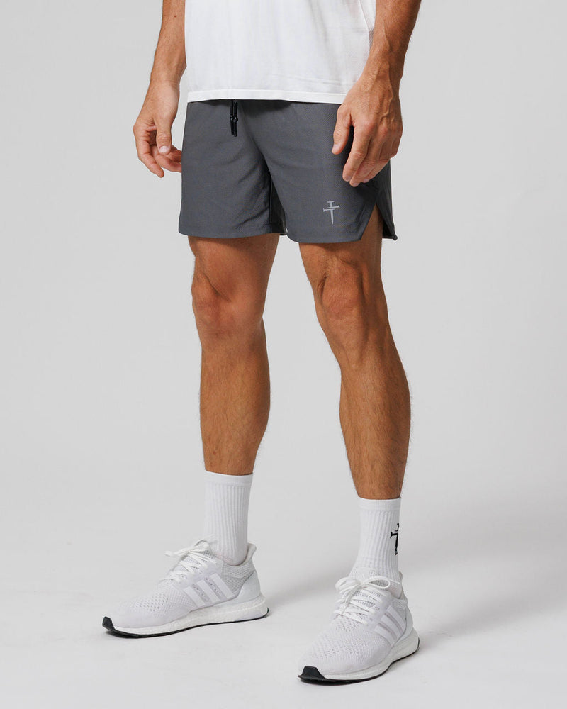 Model wearing Pro-Tech 2.0 Linerless Shorts in charcoal gray, front side angle view, paired with white Adidas sneakers.