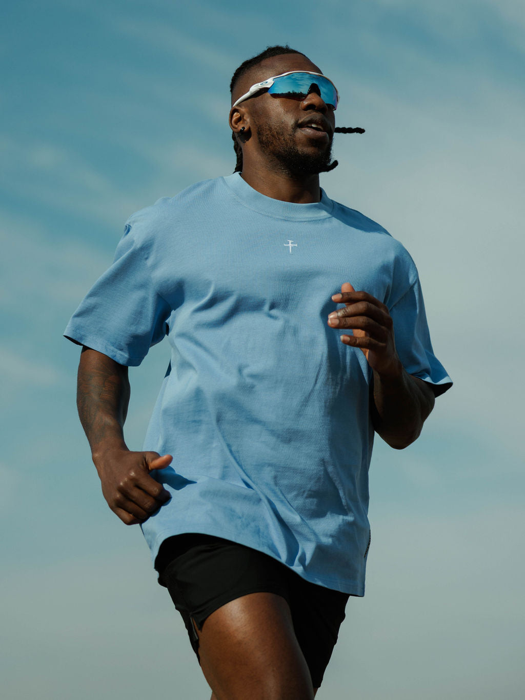 Man running outdoors wearing the Heavyweight Cotton Oversized Tee in Sky Blue, front view with small cross logo visible.