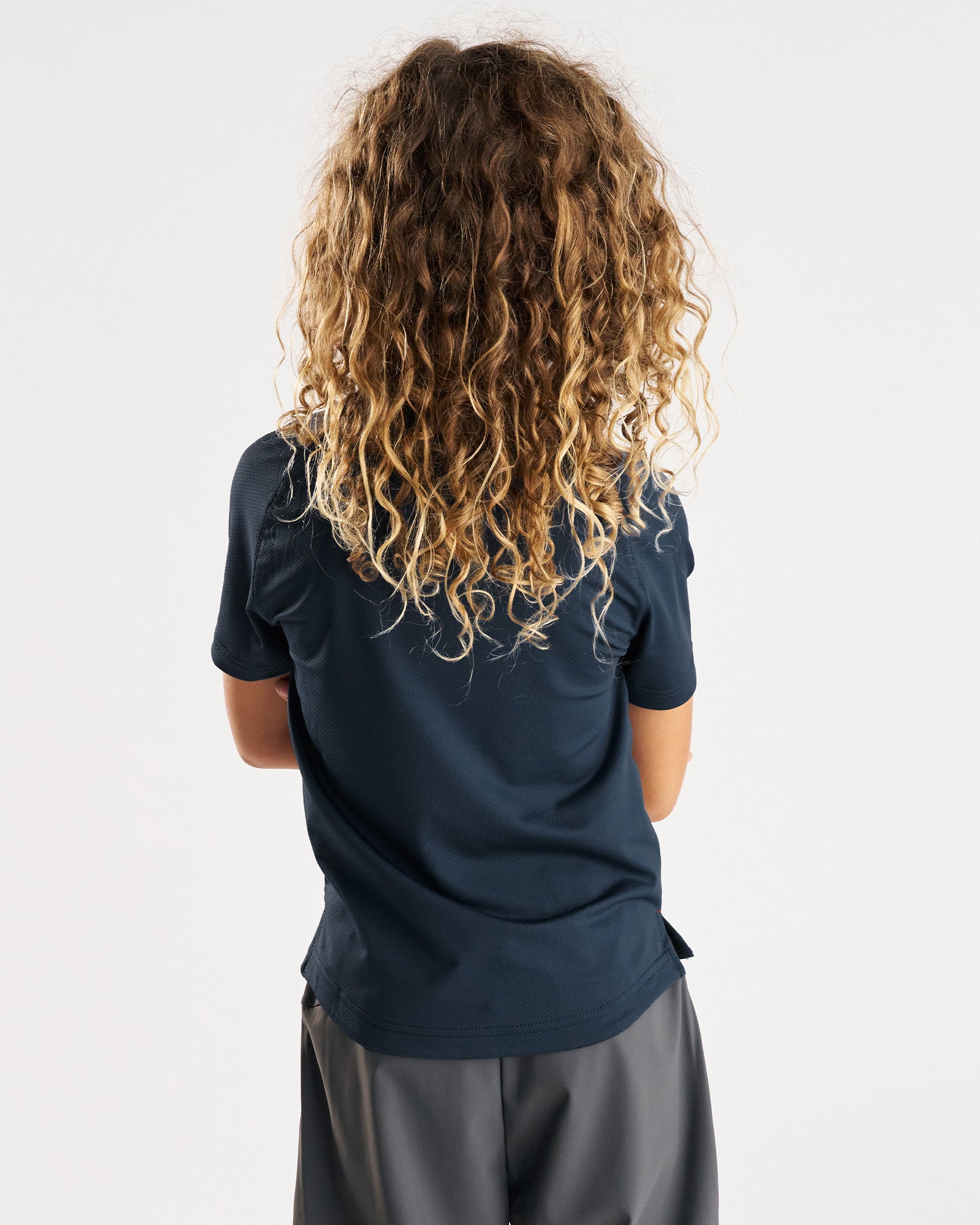 Youth Pro-Tech Tee Cross in Midnight Navy shown from the back on a young model with curly hair, paired with gray shorts.